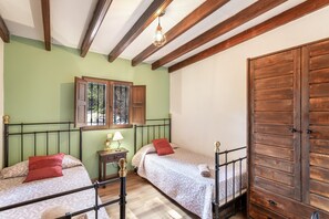 A room with two beds, wooden beams, a window with shutters, a small table with a lamp, and a large wooden wardrobe.
