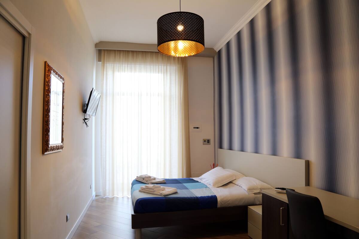 A modern hotel room with a bed, a desk, and a large window with curtains.