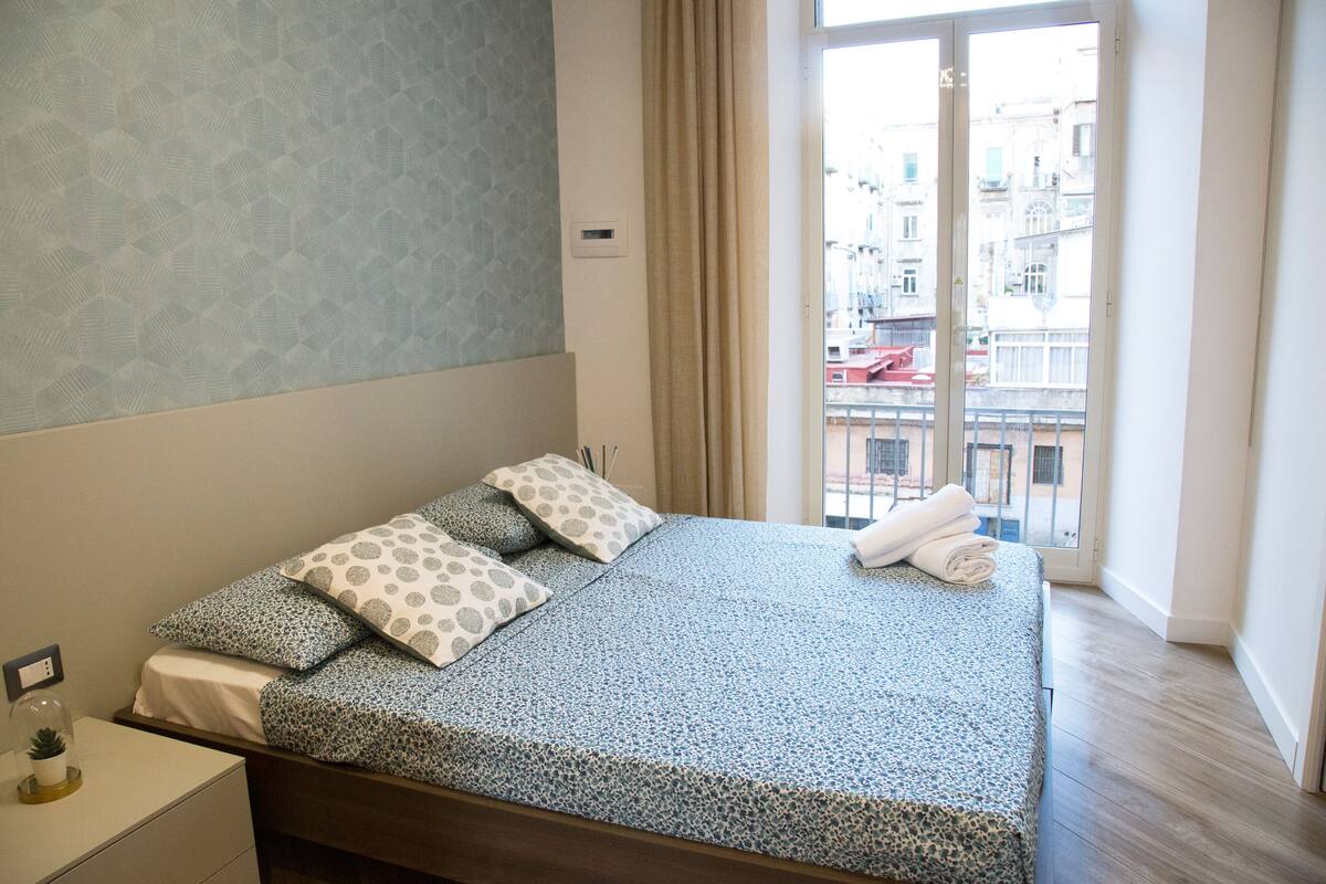 A neatly made bed with patterned bedding, a bedside table with a lamp, and a window with a view of buildings.