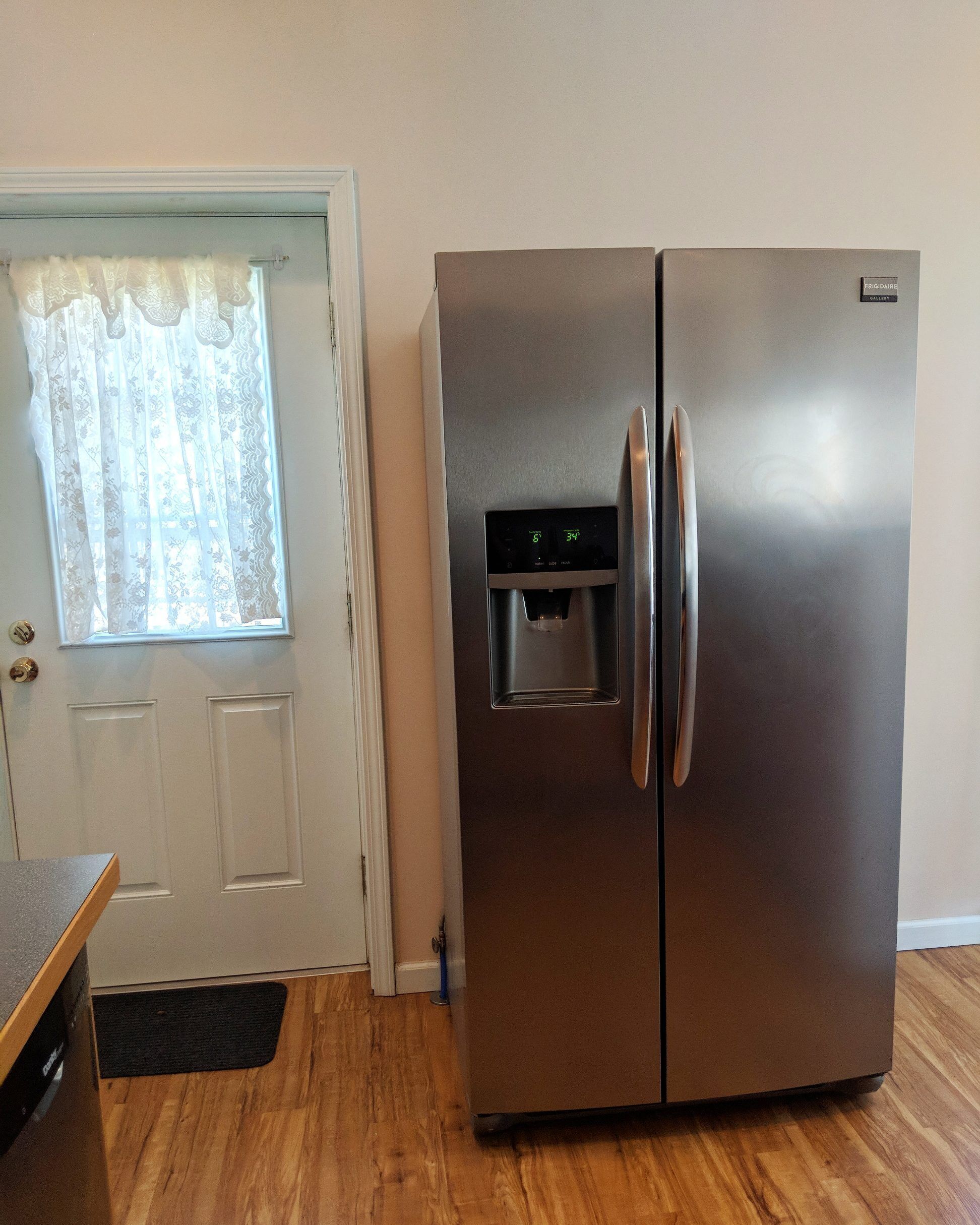 A stainless steel refrigerator with a water dispenser, a wooden floor, and a white door with a window.