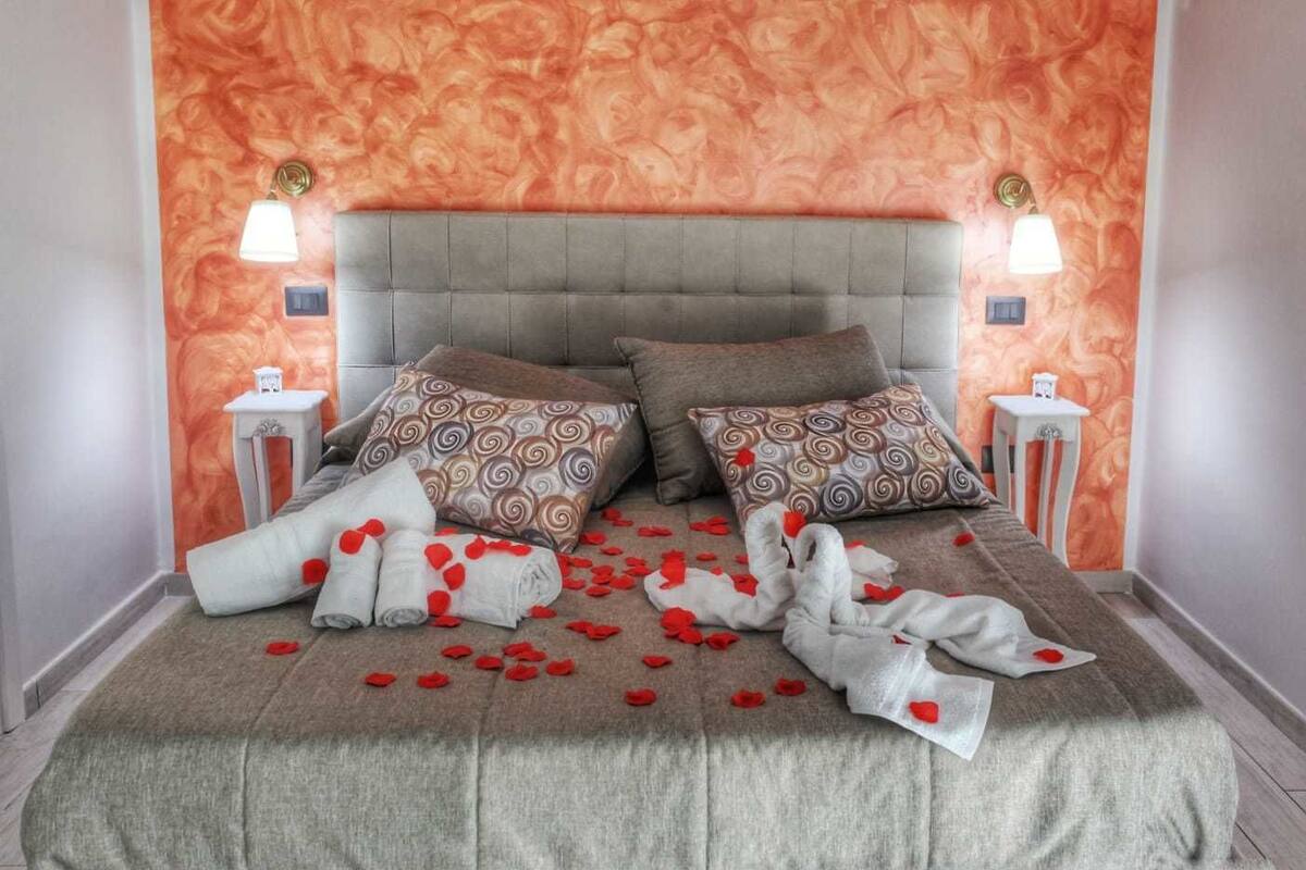 A bed with a grey headboard, two patterned pillows, and a blanket with red rose petals. There are two bedside tables with lamps and a small table with a decorative item.