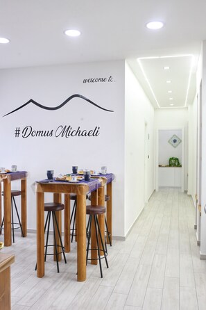 Un comedor moderno con mesas y taburetes de madera, un letrero de bienvenida y un pasillo que conduce a una habitación con una planta decorativa.