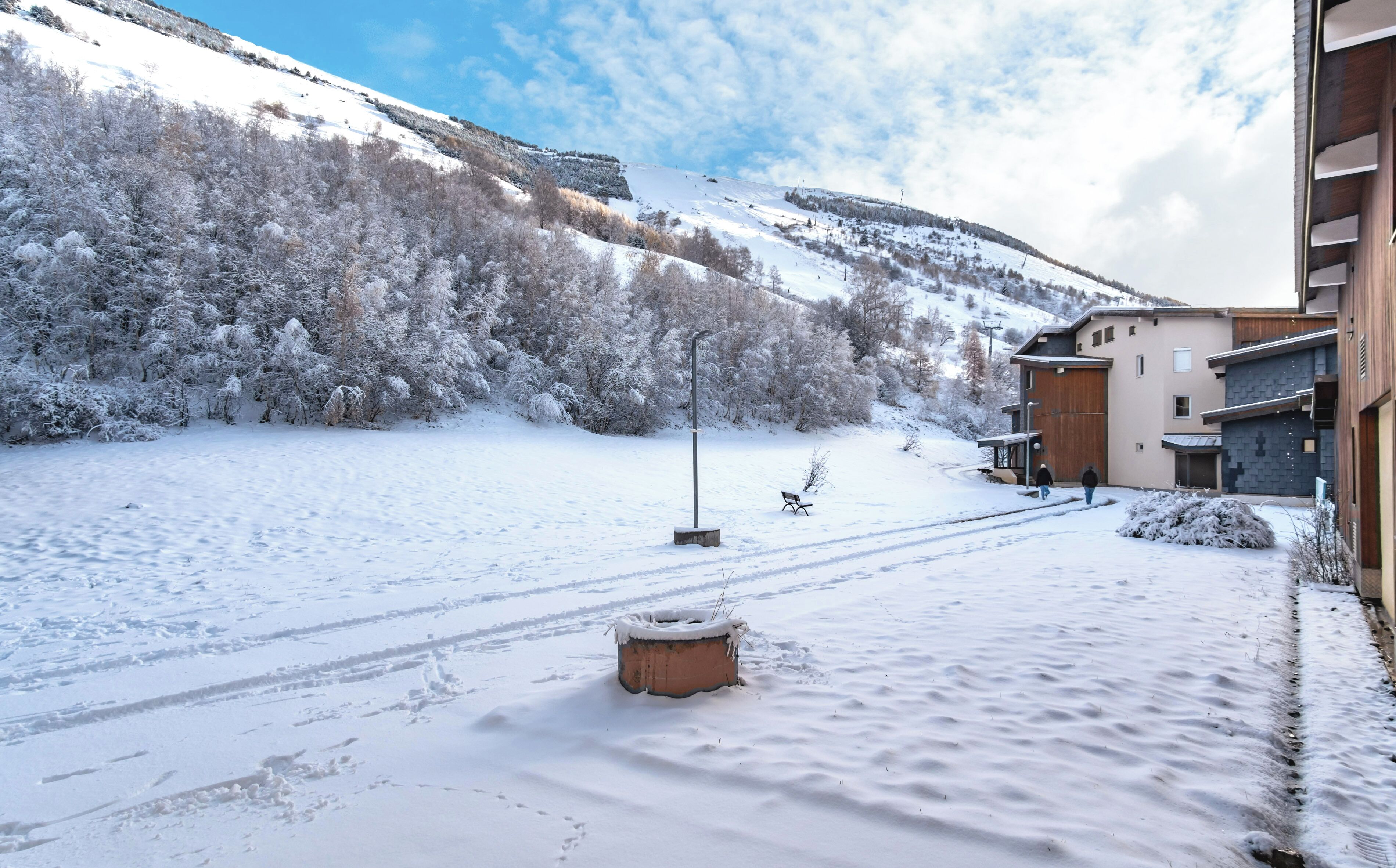 Un espace extérieur enneigé avec un banc, un poteau recouvert de neige et un bâtiment doté d’un toit en pente.
