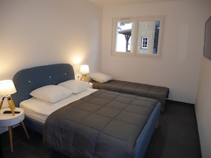 A bedroom with two beds, a nightstand with a lamp, and a window with a view of a building.