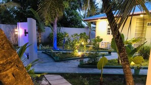 Un espacio exterior bien iluminado con un camino que lleva a una zona de estar, rodeado de abundante vegetación y un edificio con fachada amarilla.