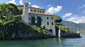 A historic building with a tower, situated by a lake with a mountainous backdrop. - Como Lake view from olive garden, balcony and pool (Tremezzina)
