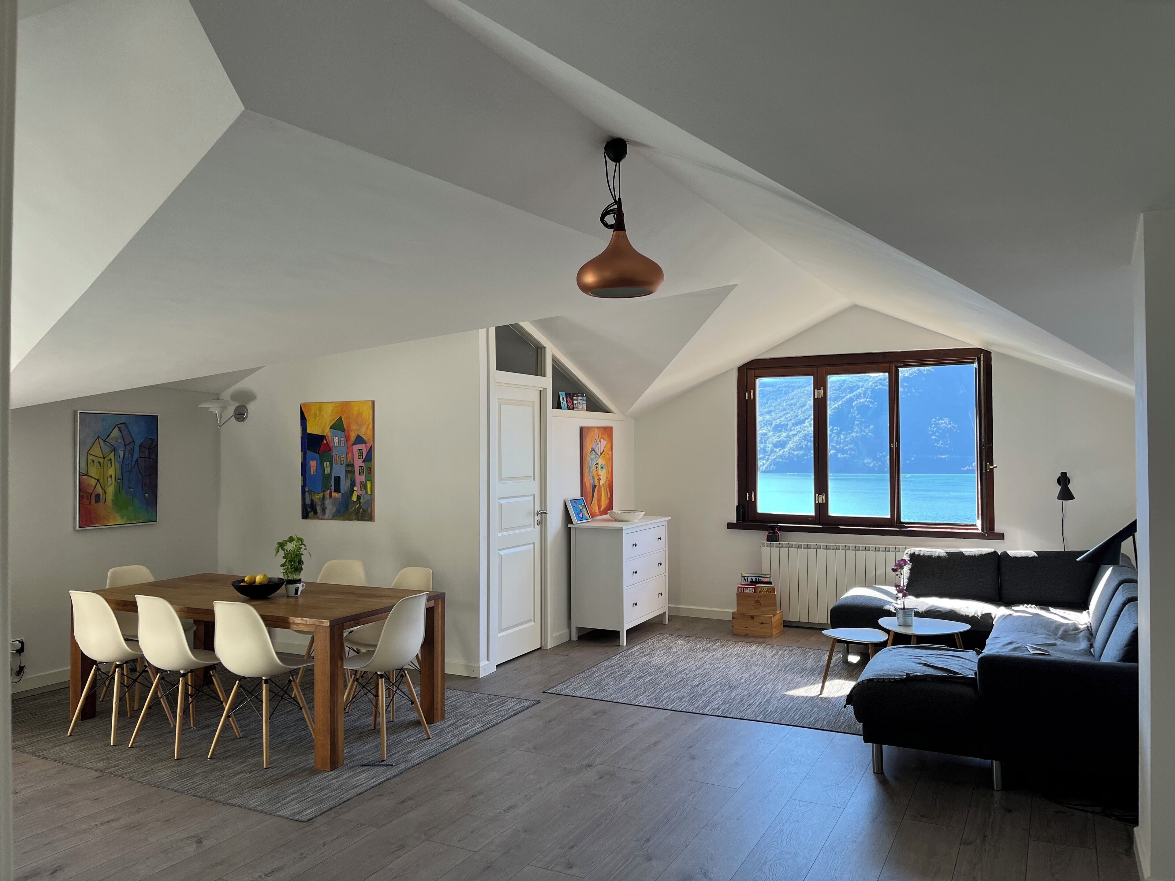 A spacious loft with a dining area, a sofa, and a large window overlooking the sea.