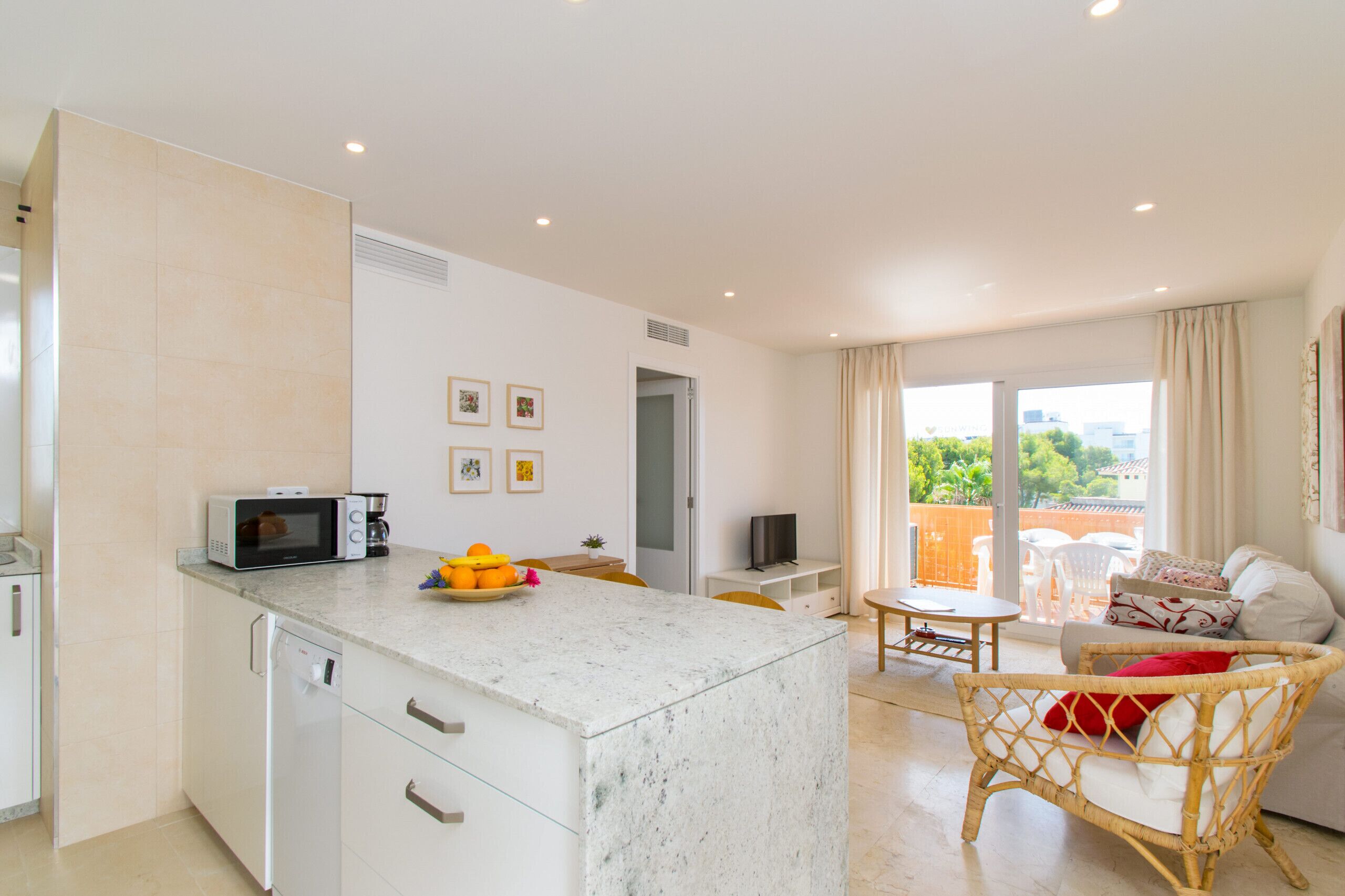 A modern kitchen with a microwave, coffee maker, and fruit bowl. There is a dining area with a table and chairs, and a wicker chair with a cushion. A television is on a stand, and a sliding glass door leads to an outdoor area with a view of greenery.