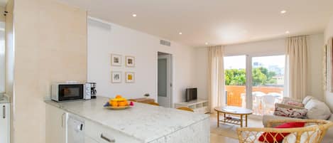 A modern kitchen with a microwave, coffee maker, and fruit bowl. There is a dining area with a table and chairs, and a wicker chair with a cushion. A television is on a stand, and a sliding glass door leads to an outdoor area with a view of greenery.