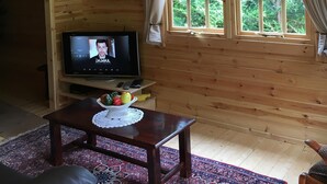 Interior de uma cabana de madeira com televisão, uma mesa com um cesto de frutas, um tapete estampado e uma janela com cortinas.