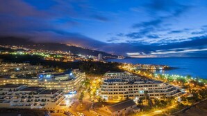 Paisagem costeira urbana à noite, com edifícios iluminados, um porto de iates e vista para o mar.