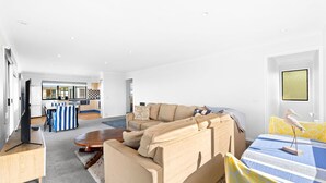 A modern living room with a sofa, coffee table, and a dining area with a table and chairs. - 45 Hillside Cres Beach House (Kianga)