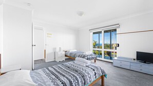A modern bedroom with two beds, a TV, and a sliding glass door leading to a balcony. - 45 Hillside Cres Beach House (Kianga)