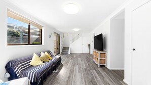A modern, well-lit room with a sofa set, a television on a wooden cabinet, and a staircase leading to another level. - 45 Hillside Cres Beach House (Kianga)