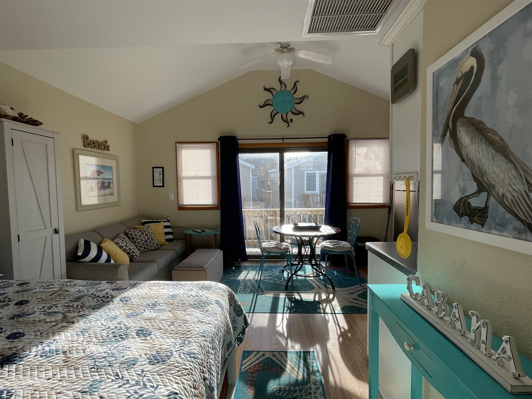 A cozy room with a bed, a sofa, a dining table with chairs, a ceiling fan, and a large window with curtains.