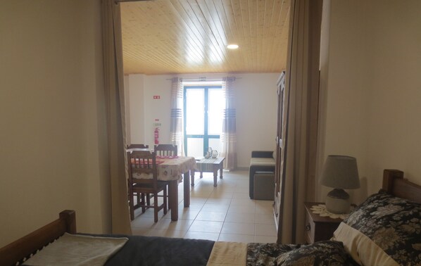 A room with a bed, a table, and a sofa. There is a window with curtains, a door, and a lamp on a bedside table.