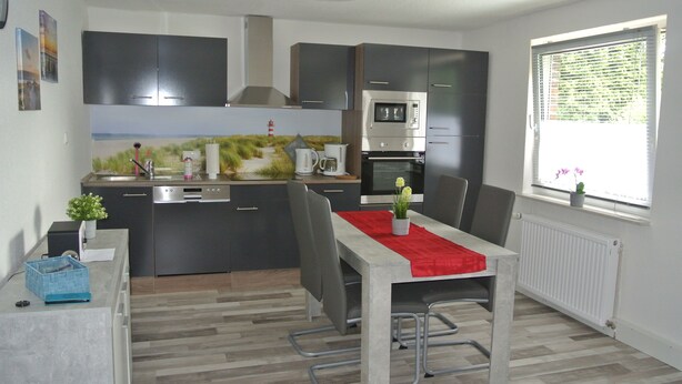 A modern kitchen with dark cabinets, a built-in oven, and a dining area with a red table runner. - Holidays, holidays and sea (Wilhelmshaven)
