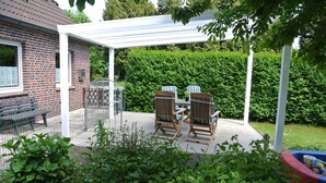A covered patio with two wooden chairs and a table, a brick house, and a hedge in the background. - Holidays, holidays and sea (Wilhelmshaven)