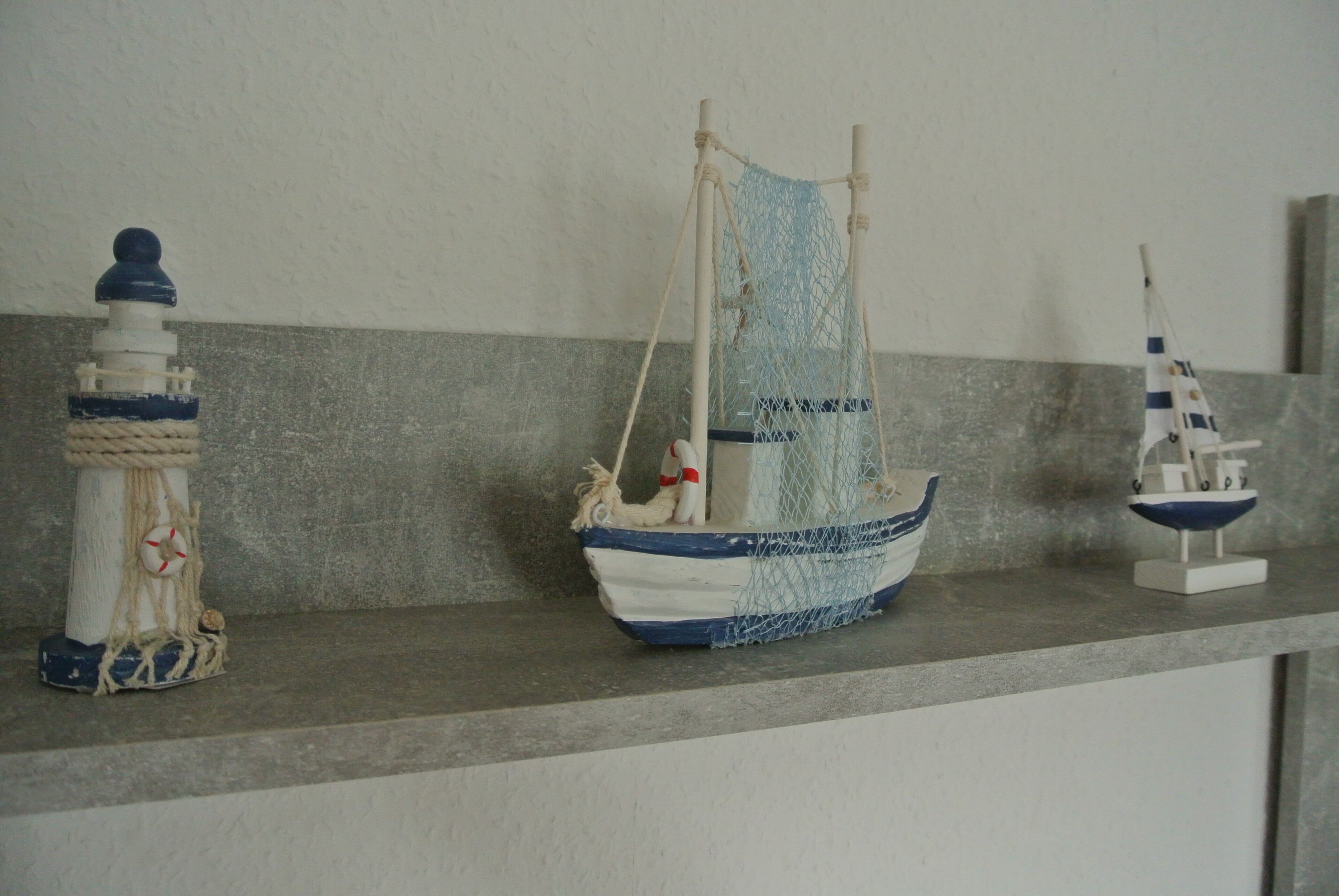 Une étagère ornée de trois objets décoratifs sur le thème de la marine : un phare, un voilier et une maquette de petit navire.