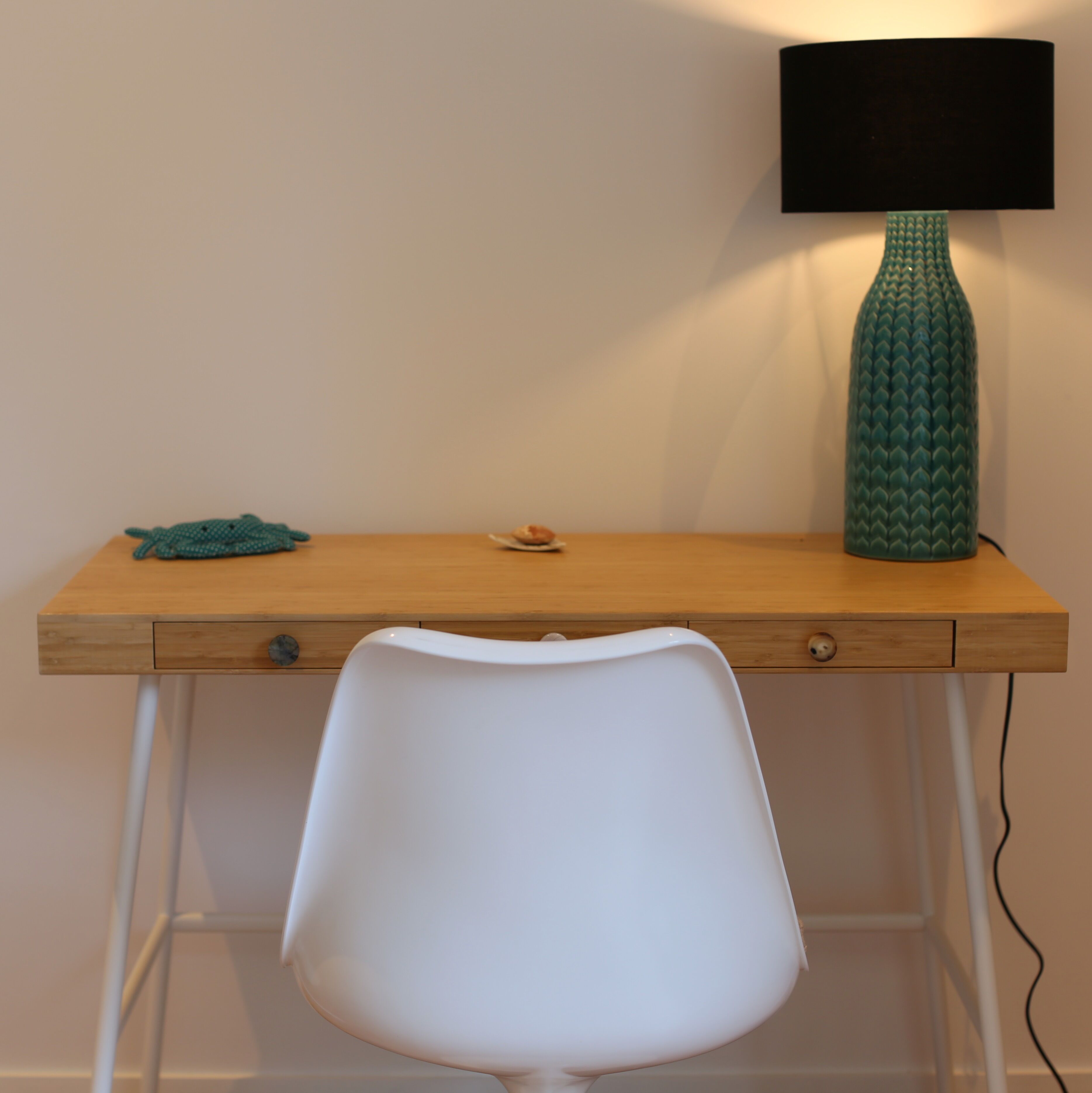 A wooden desk with a white chair, a teal vase, and a black lamp.