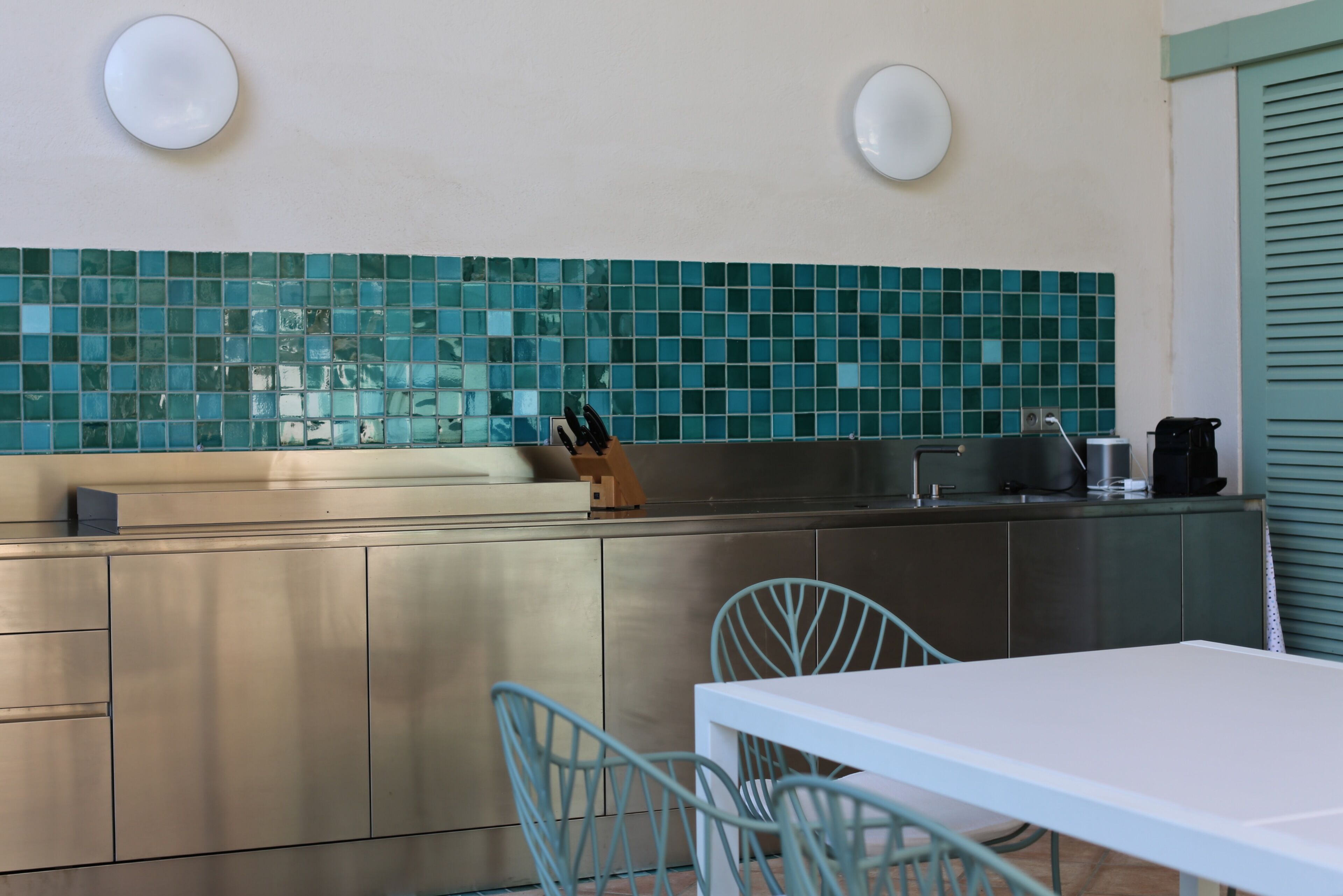 A modern kitchen with teal mosaic backsplash, stainless steel cabinets, and a white dining table with blue chairs.