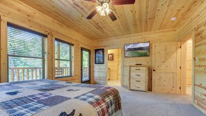 Ein Schlafzimmer einer Holzhütte mit einem großen Bett, einem Deckenventilator, einem Fernseher und einer Tür zu einem anderen Raum.