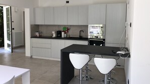 A modern kitchen with white cabinets, a black countertop, and a built-in oven. There are two white bar stools and a white table. - VILLA GINEVRA COLICO (Colico Piano)