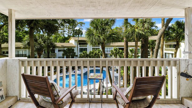 A balcony with two wooden chairs, a small table, and a view of a swimming pool and palm trees. - Reduced Rates! Short Walk to the Beach and Pool! (Hilton Head Island)
