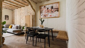 A dining area with a rustic stone wall, a wooden ceiling, and a painting of a dining scene. There is a sofa and a table set with chairs. - CASA CARME SEGUNDO (Barcelona)