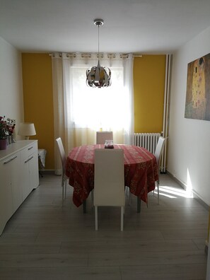 A dining area with a round table covered in a red tablecloth, white chairs, a chandelier, and a window with curtains.