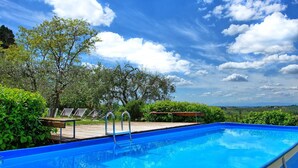 Piscina azul límpida com um deque de madeira e cadeiras de descanso, rodeada por vegetação exuberante e árvores, sob um céu parcialmente nublado.