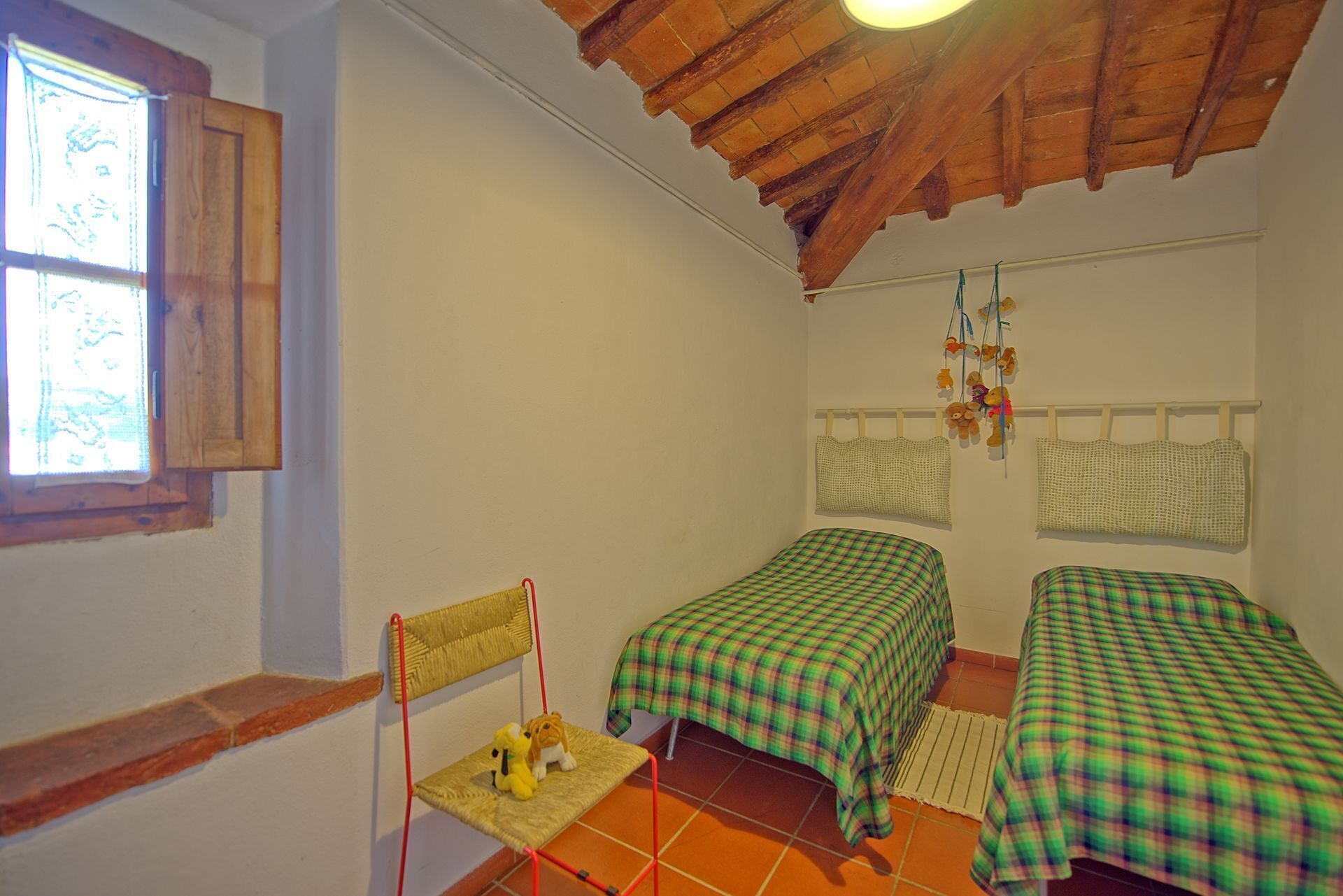 Una habitación con dos camas, una silla pequeña, un ventanal y un techo con vigas a la vista.