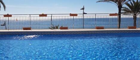 A swimming pool with clear blue water, surrounded by a fence and palm trees, with a view of the sea in the background.