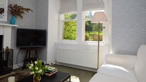 A living room with a fireplace, a television, a window with a view of greenery, and a white sofa. - Garden cottage, estate property set in amazing gardens, pet friendly, Wi-Fi (Strachur)
