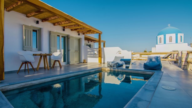 A pool area with a wooden pergola, white walls, and a blue-domed building in the background. - Aeris suites (Naxos)