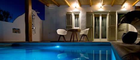 A swimming pool with a wooden pergola overhead, two white chairs, and a door with glass panels.