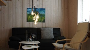 A cozy room with a wooden ceiling, a black sofa, a wooden chair, a small table, and a painting on the wall.