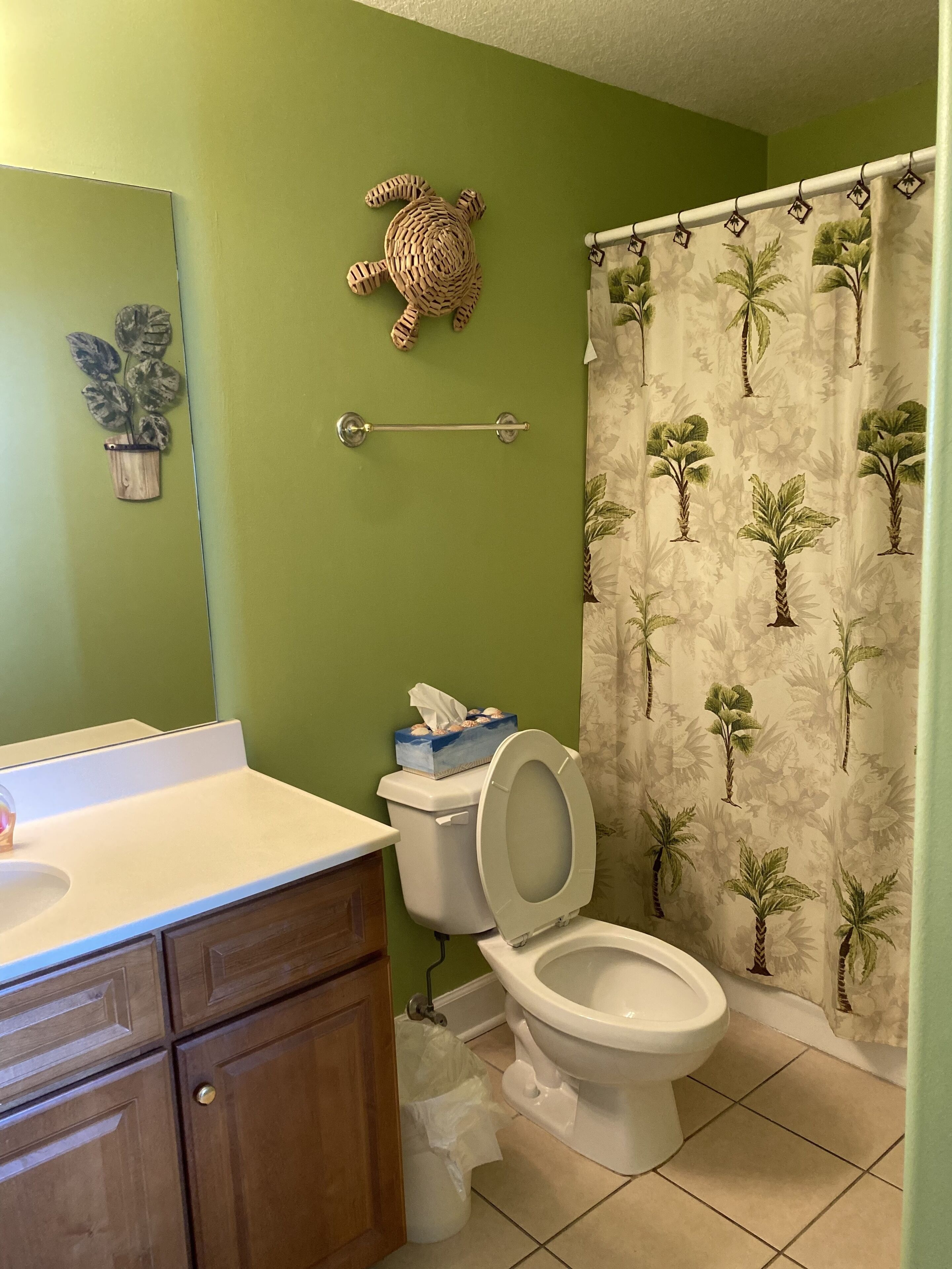 Une salle de bain avec un mur vert, une décoration en forme de tortue en bois, des toilettes avec un porte-mouchoirs et un rideau de douche orné de motifs de palmiers.