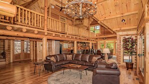 A spacious log cabin interior with a large wooden ceiling, a central seating area with a sofa and coffee table, and a balcony with a railing.