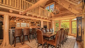 A spacious wooden cabin interior with a large dining table, multiple chairs, a kitchen area, and a staircase leading to a loft.