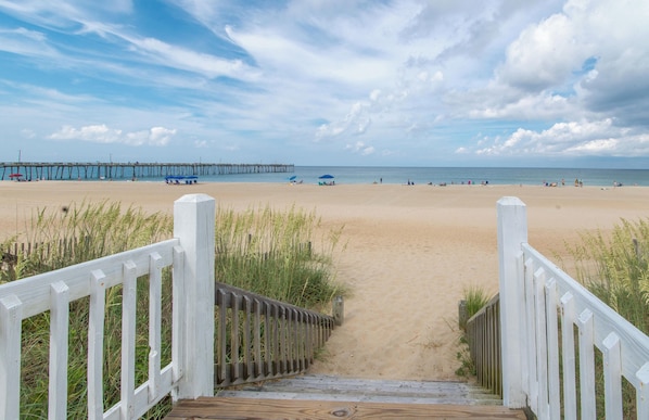A wooden boardwalk leading to a sandy beach with a pier in the distance.