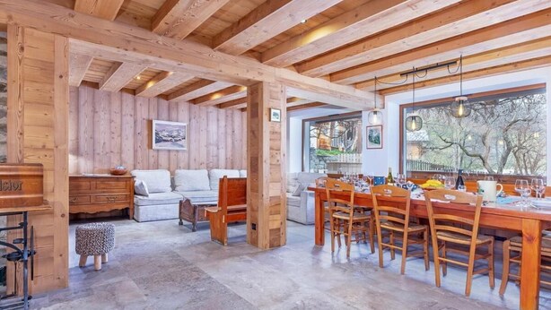 A spacious room with a wooden ceiling, a dining area with a long table and chairs, a sofa, and a small wooden cabinet.