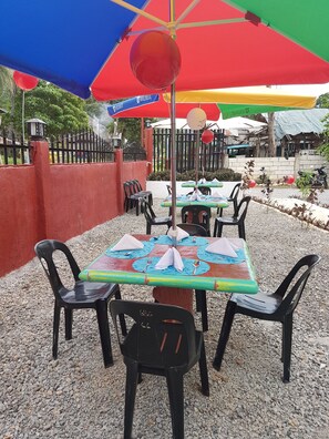 Un área de comedor al aire libre con mesas, sillas, sombrillas y una cerca roja.