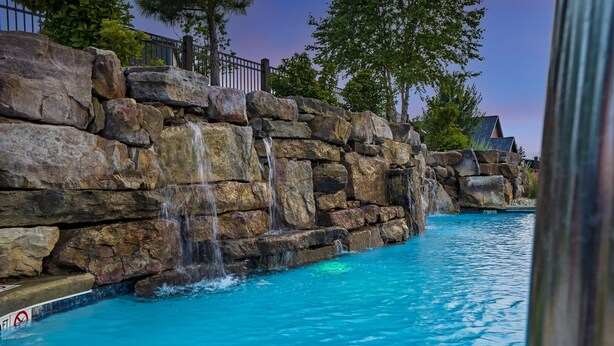A swimming pool with a rocky waterfall feature, surrounded by trees and a fence.