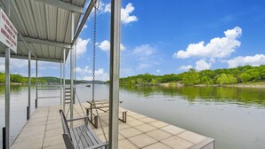 A covered dock with a swing chair and a bench, overlooking a calm lake with a pier in the distance.