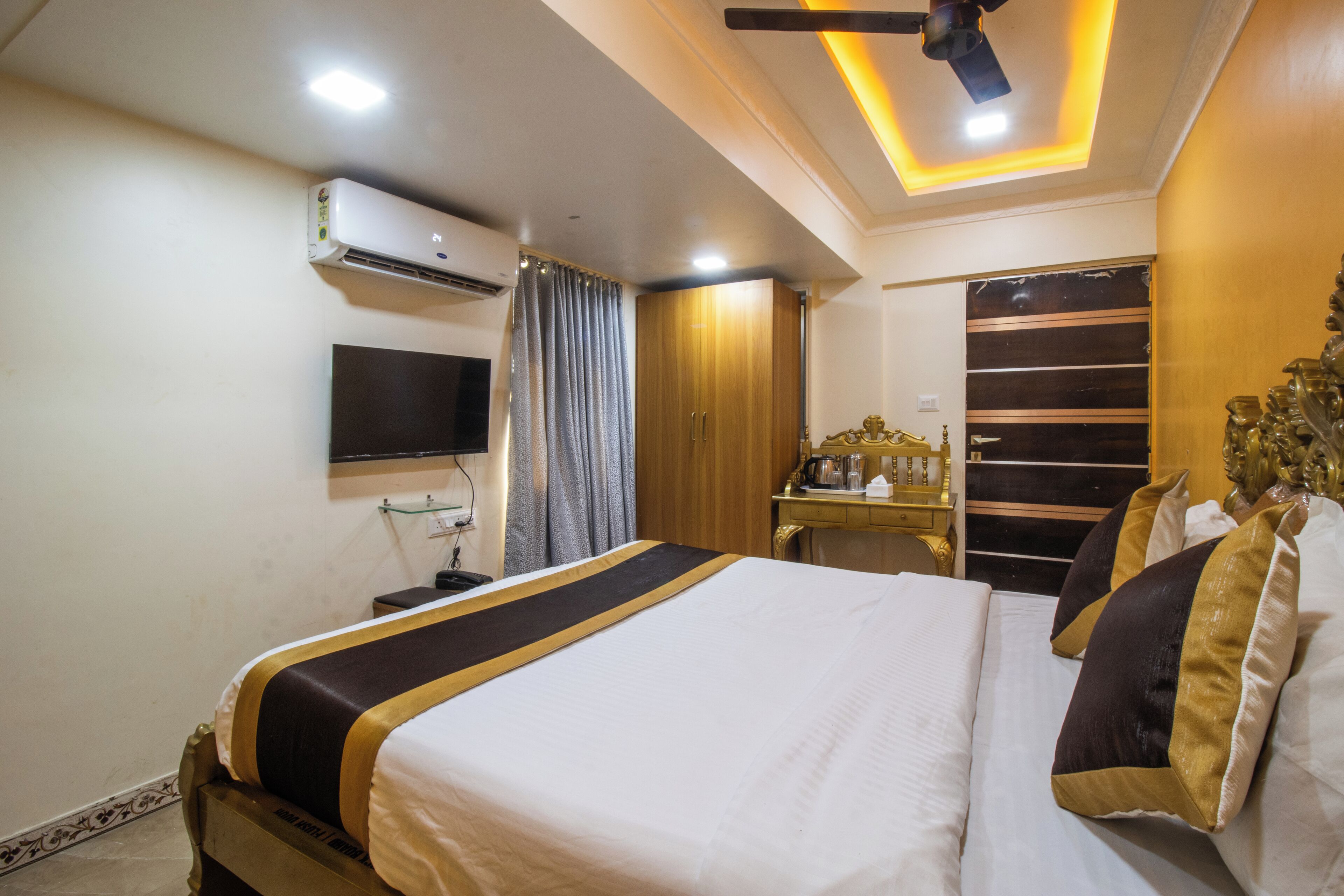 A hotel room with a bed, a TV mounted on the wall, an air conditioning unit, a desk with a chair, and a decorative item on the wall.