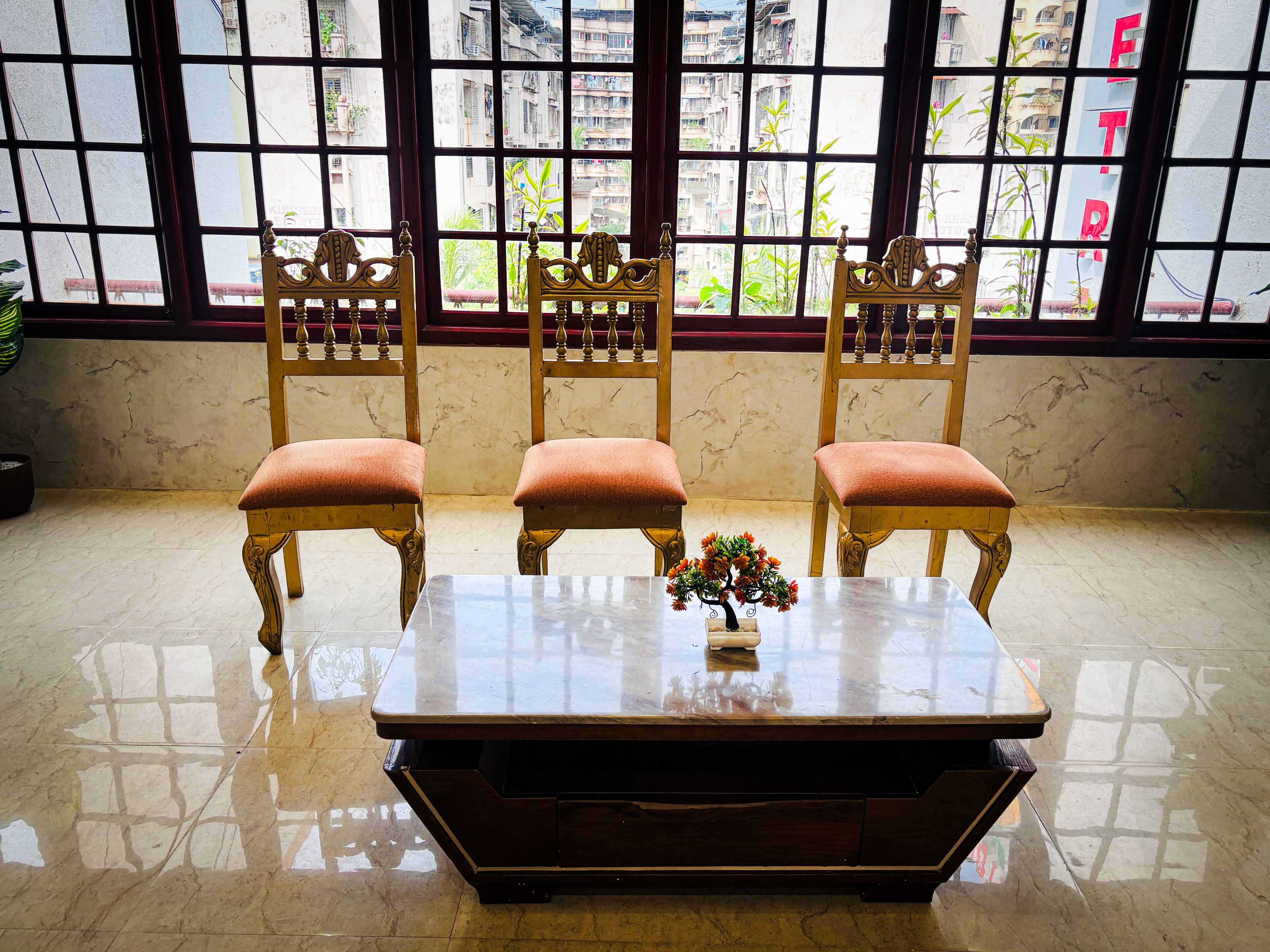 A room with a marble floor, a wooden table with a vase of flowers, and three wooden chairs with cushions.