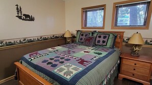 A bedroom with a wooden bed, two bedside tables with lamps, a quilt, and two windows with blinds.