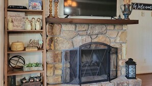 A living room with a stone fireplace, a television mounted above it, and a wooden shelf with various decorative items.
