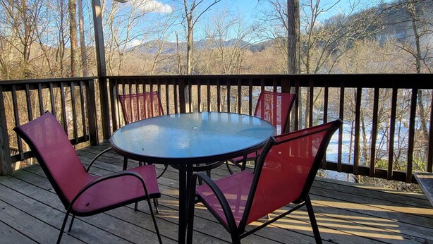 A wooden deck with a round table and four chairs, overlooking a body of water and trees.